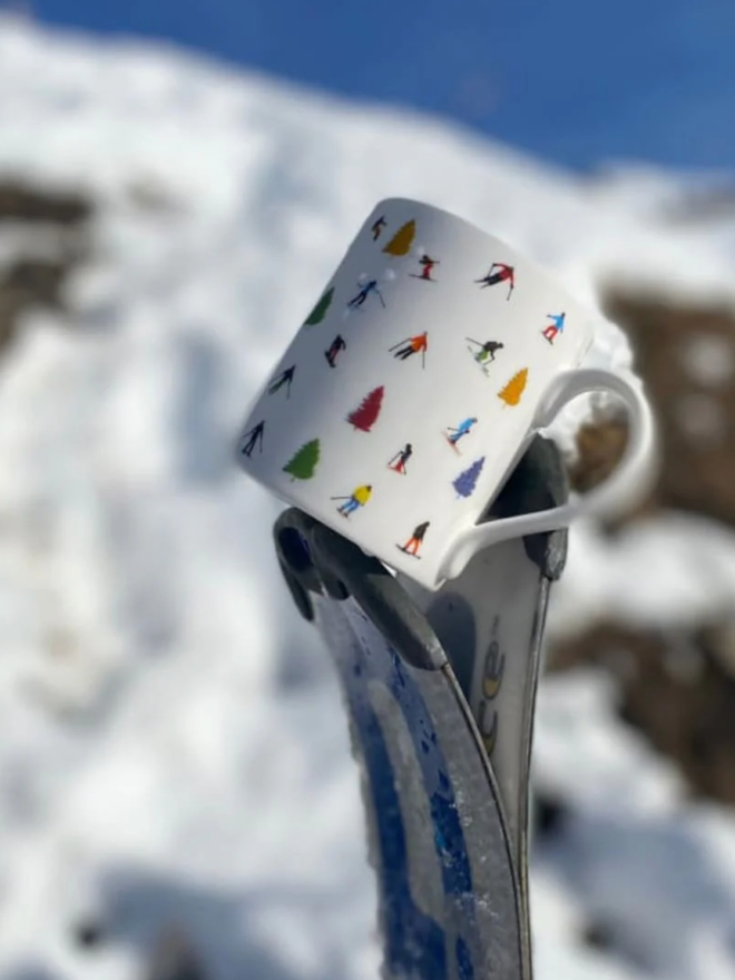 Tree Skiing Mug. A mug held by a dark grey support, against the backdrop of a snowy mountain. The mug is a simple white with trees and skiers illustrated on it.