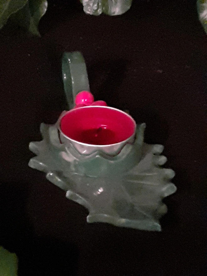 Front view of a handmade ceramic Christmas candle holder shaped like a holly leaf with a red tealight and loop handle.