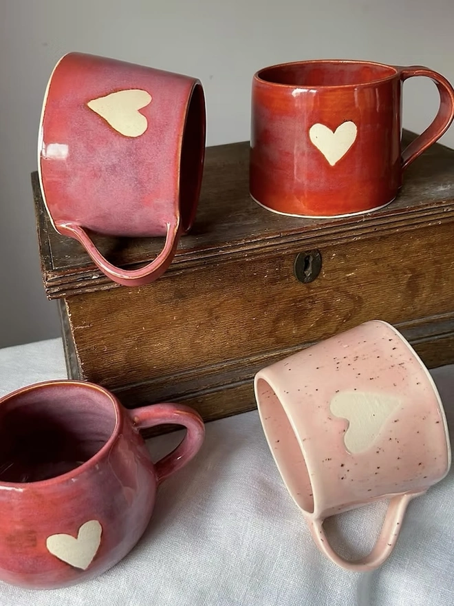 Heart Mug Strawberry Pink, a mug on a white surface in front of more red heart mugs, on a small wooden cabinet. 