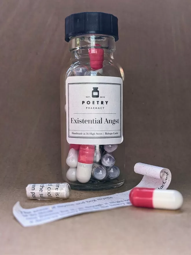 A glass bottle of Existential Angst poetry pills against a brown paper background.  in front of the bottle there are two pills, one red and white, one clear with visible text on the paper inside it, and a slip of rolled paper with a poetry quote printed on it