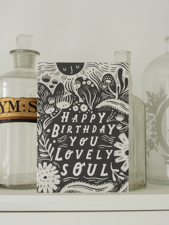 Black and white greeting card with illustration and the words Happy Birthday you lovely soul written on it stood up on a shelf with glass medicine bottles behind it 