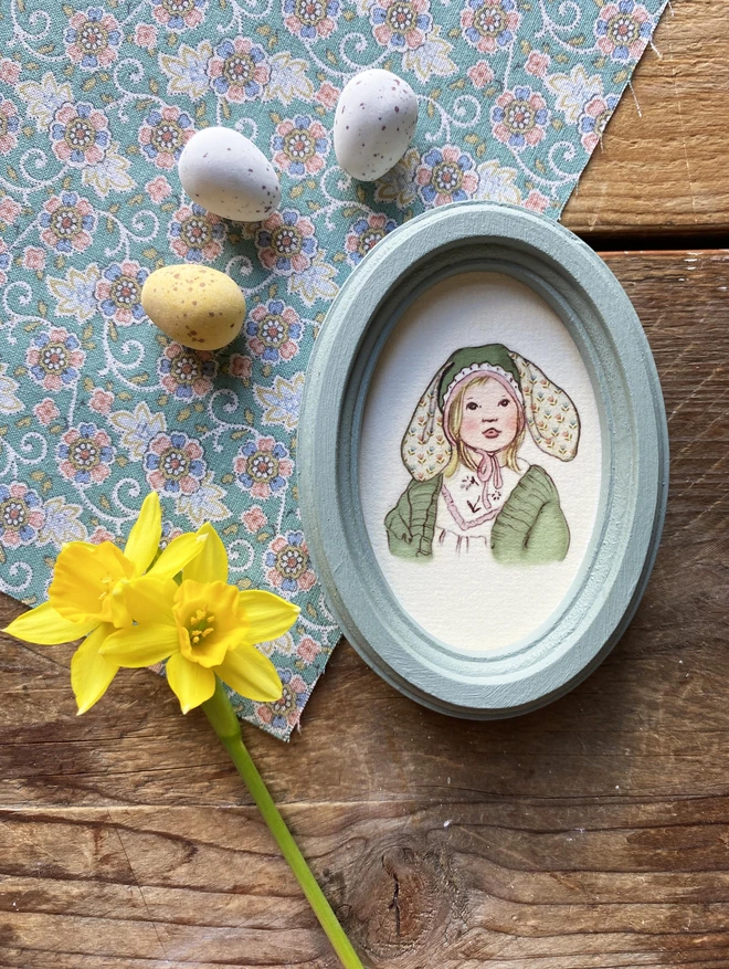 a photograph of a miniature portrait of a little blonde girl wearing an easter bonnet with big bunny ears framed in a duck egg blue oval wooden frame photographed next to a daffodil and mini eggs