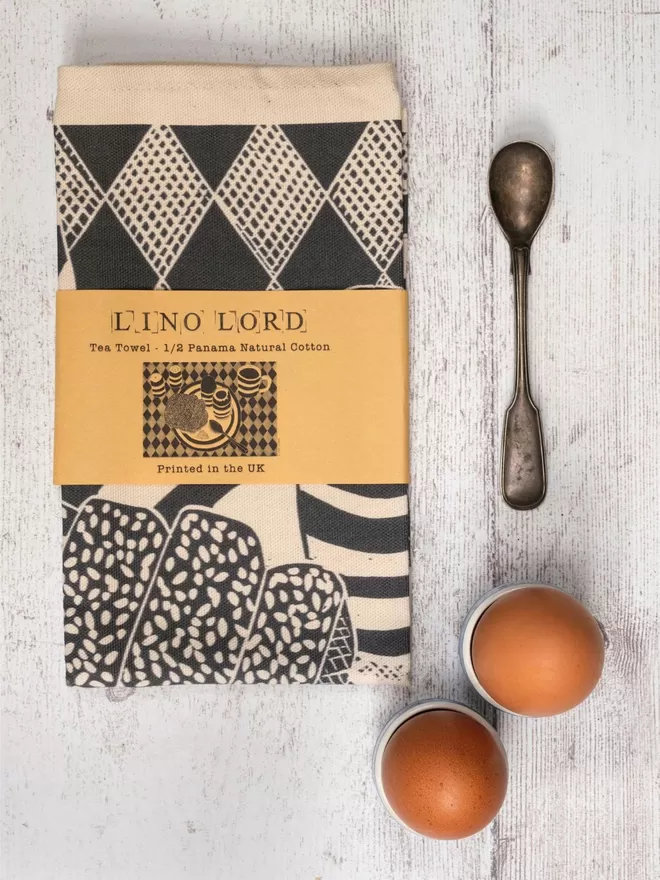 Picture of a tea towel with an image of eggs & soldiers on a plate with a drink and salt & pepper, taken from an original lino print