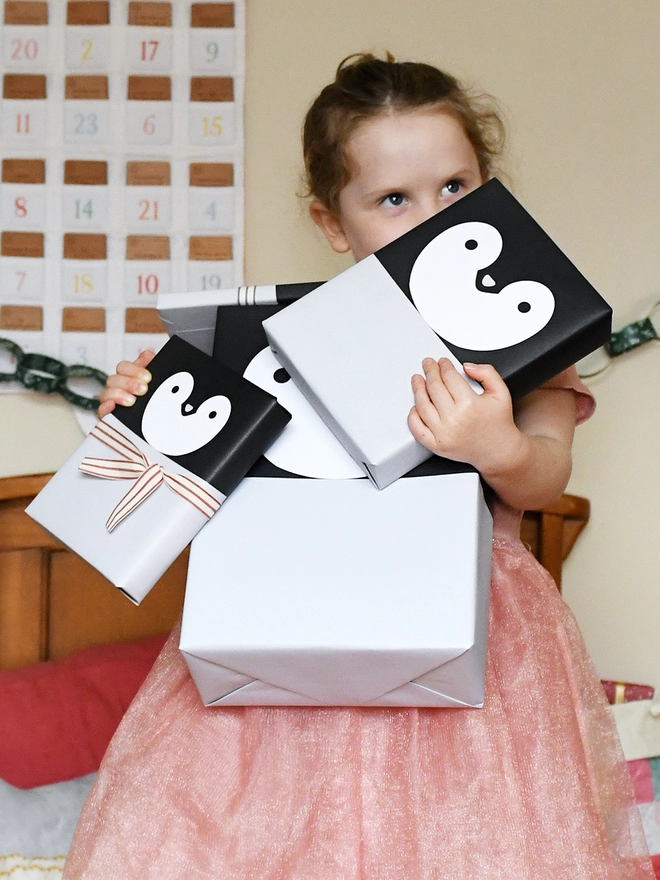 A young girl wearing a pink tutu is holding several gifts wrapped in penguin wrapping paper.