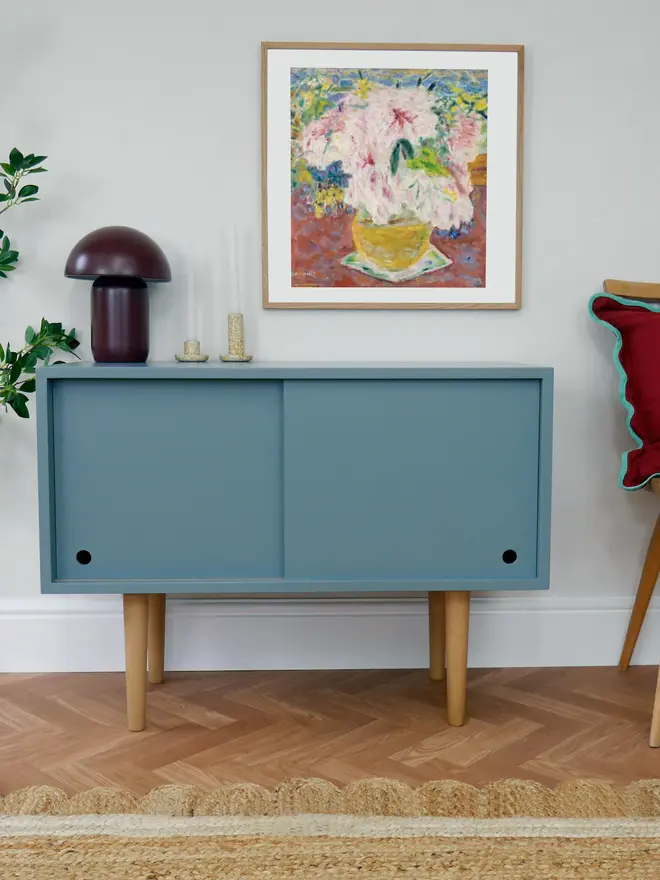 Teal mid-century cabinet with sliding doors and wooden legs in a styled room.