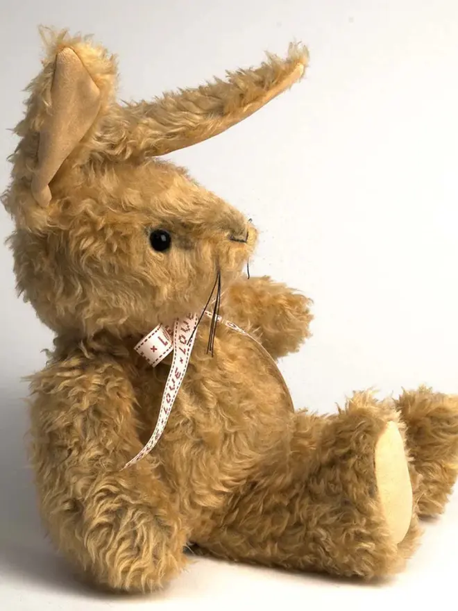 Handmade brown stuffed rabbit with long ears and a pink ribbon bow sitting upright against a plain background.