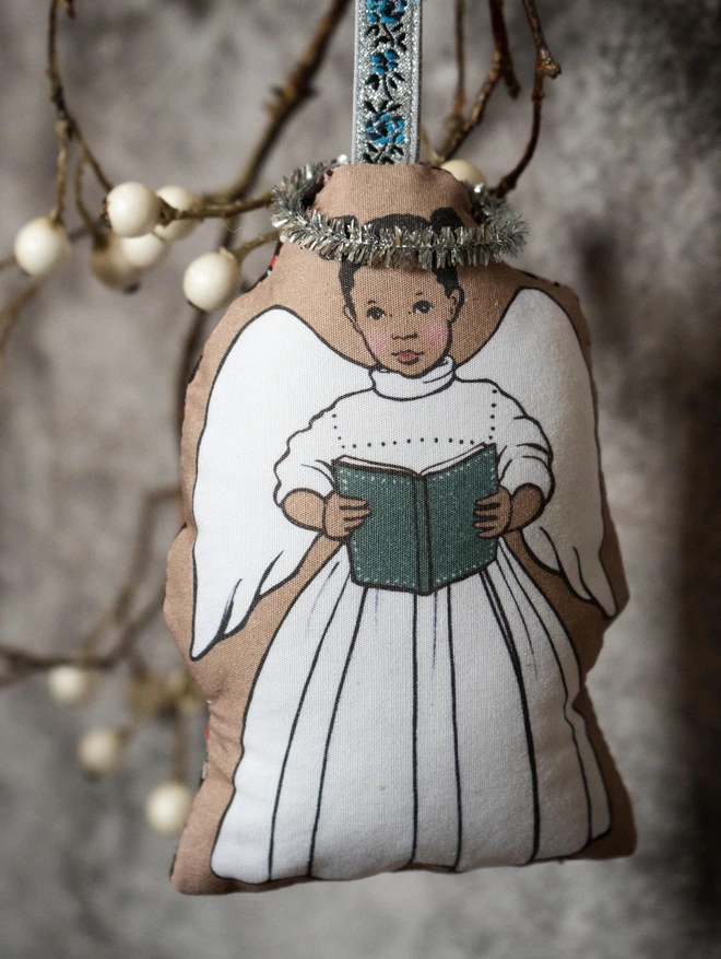 a christmas tree plush hanging angel decoration illustrated in a vintage story book style of an afro caribean girl with top knots holding a blue book