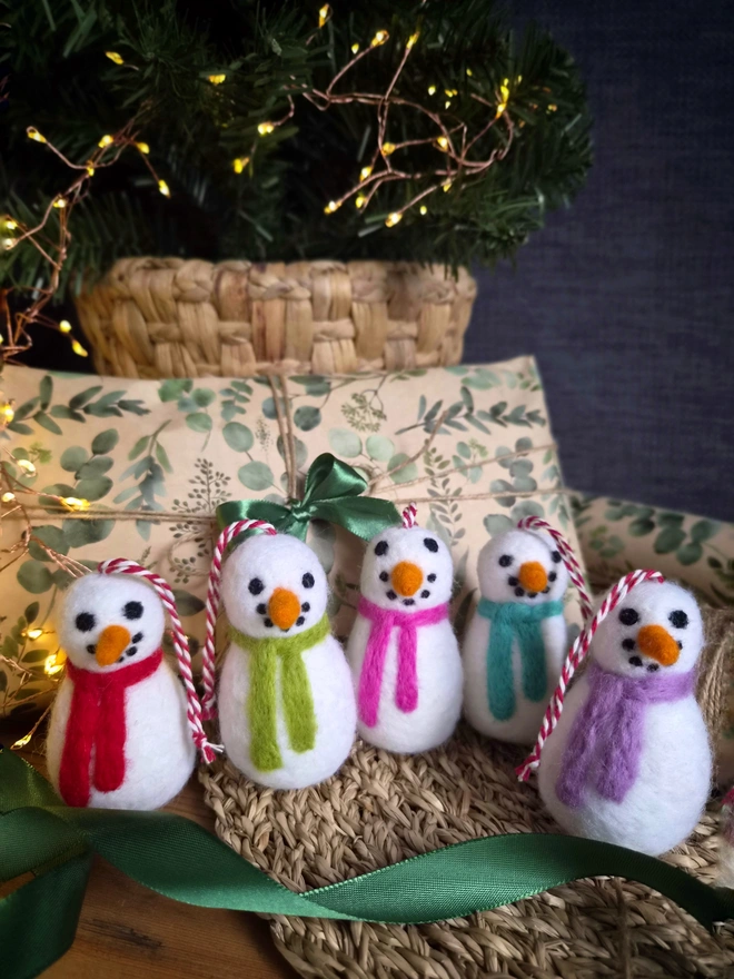 Felt snowmen decorations with red and white twine to hang from a Christmas tree. All placed in front of a Christmas tree and present.