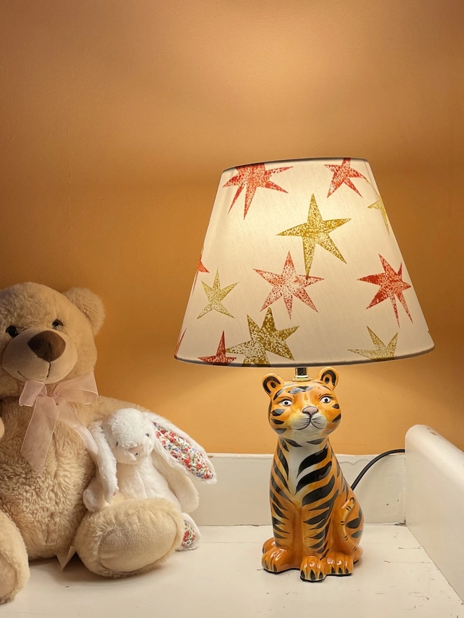 A dimly lit kids bedroom corner with a cream lampshade decorated with bright orange and yellow star patterns, styled in a kids bedroom on a tiger lamp base. A fun empire lampshade and kids bedroom decor piece that adds warmth and character.