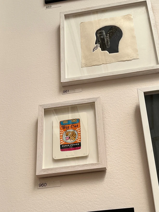 Big Cat Power Battery Screenprint, a colourful cat design against a white card. It is framed against a white wall accompanied by another framed design.  
