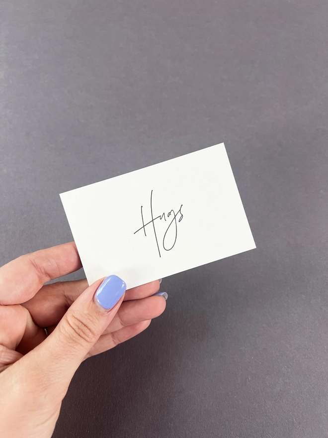 a single hug tiny note card held in a woman's hand to show scale of the cards. Hugs is letterpress printed in black ink in a modern calligraphy style