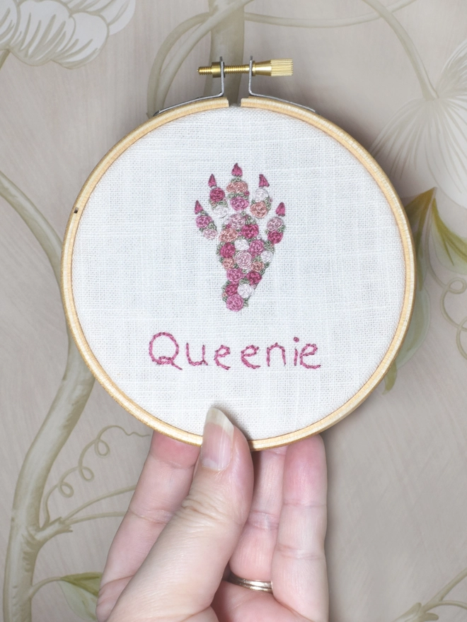 An embroidered Pink Roses Rabbit Foot, of woven wheel roses in 5 shades of pink with French Knot green grass.  Displayed in a hoop frame held up by a hand.