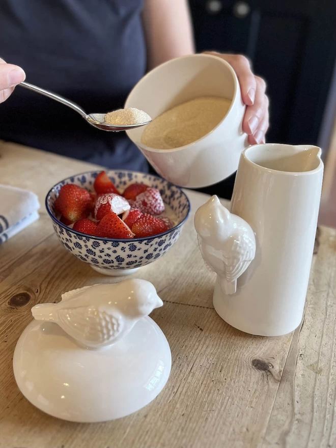 Sugar is being sprinkled over strawberries, from a handmade ceramic sugar bowl with bird lid.  