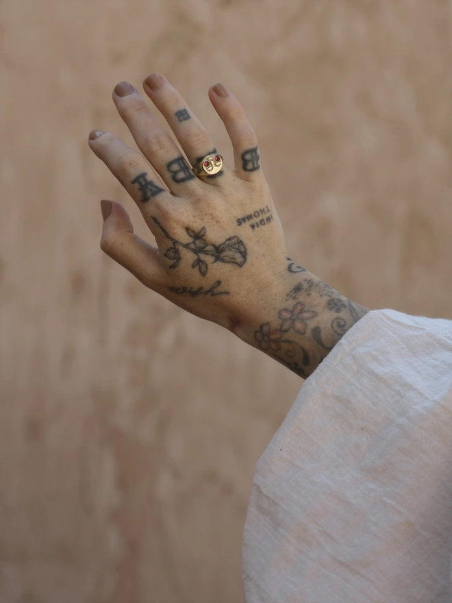 The image of a tattooed female hand wearing a moon face ring with ruby cheeks, the wearer has a puffed white fabric sleeve and the background of the image is a terracotta toned rustic plaster 