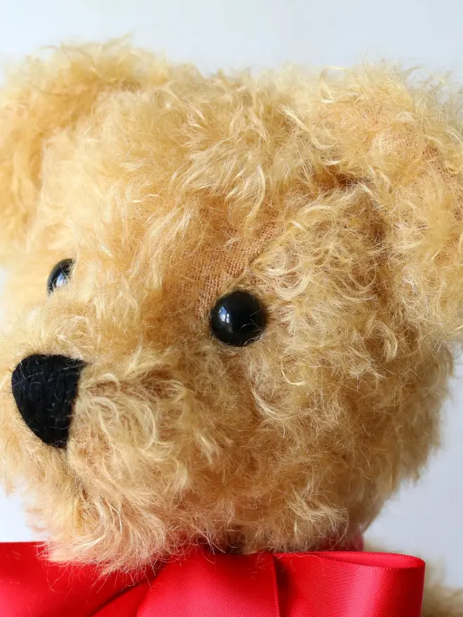 Handmade brown teddy bear sitting upright with a red ribbon bow against a plain background.