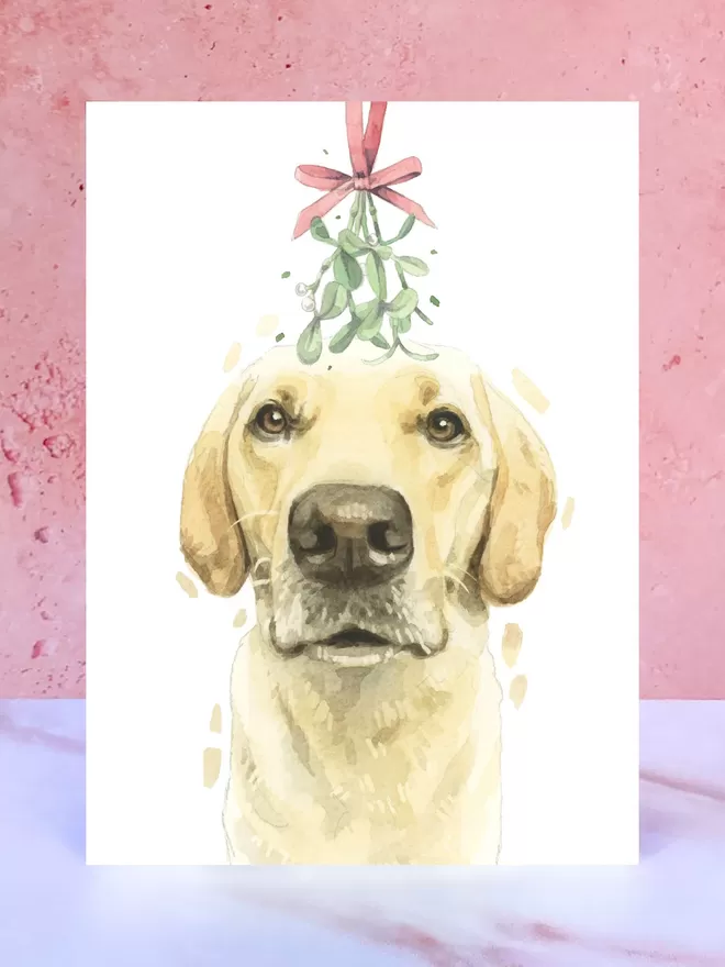 A Christmas card featuring a hand painted design of a Yellow Labrador, stood upright on a marble surface.