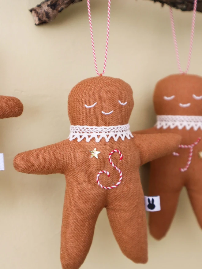 Brown linen gingerbread man christmas tree ornament with a letter hand embroidered in candy cane colours, star shaped sequin, crochet cotton lace around the neck, smile embroidered in white and candy cane colour string to hang. 