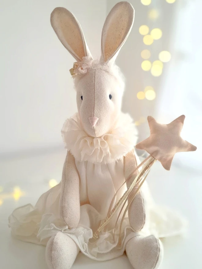 Veronica Christmas angel bunny. Front view of seated bunny wearing ivory floaty dress and choir collar, holding a pale gold satin twirly star wand.