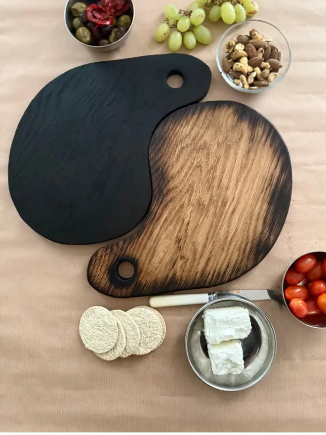 “Pair of handmade Yaki Sugi serving boards in blackened and natural oak, styled together with olives, cherry tomatoes, grapes, crackers and cheese on a rustic wooden table.”