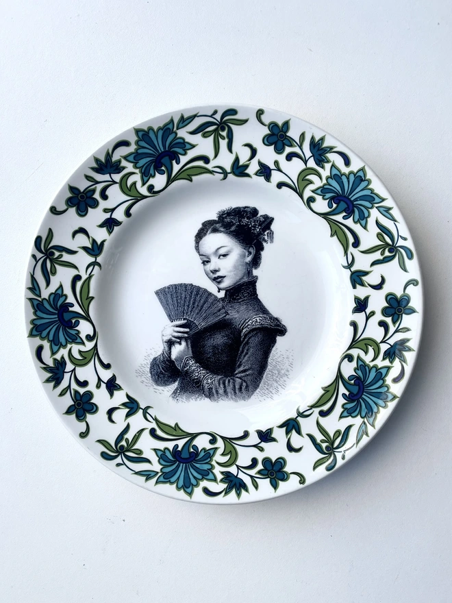a vintage plate with an ornate border has a black and white vintage illustration of a beautiful Asian girl holding a fan  in the centre