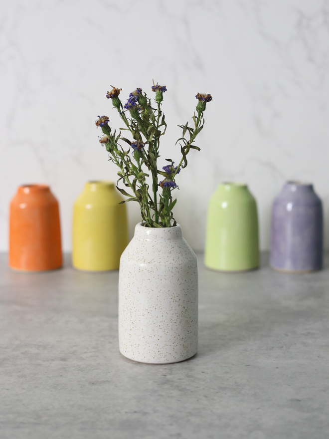 A speckled white ceramic bud vase with dried purple wildflowers in the foreground, with four handmade bud vases in orange, yellow, spring green, and lavender arranged behind it, all set on a grey marble surface against a white marble background