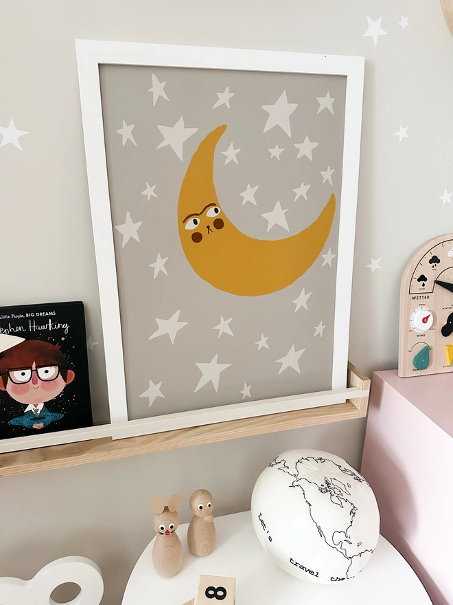 Dreamy moon print with a quirky face surrounded by stars. A celestial, modern Scandinavian style print on a shelf in a kids playroom, nursery or bedroom