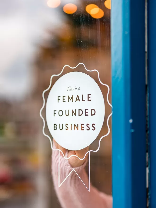 Female Founded Business Rosette Sticker seen on a shop door with a blue frame.