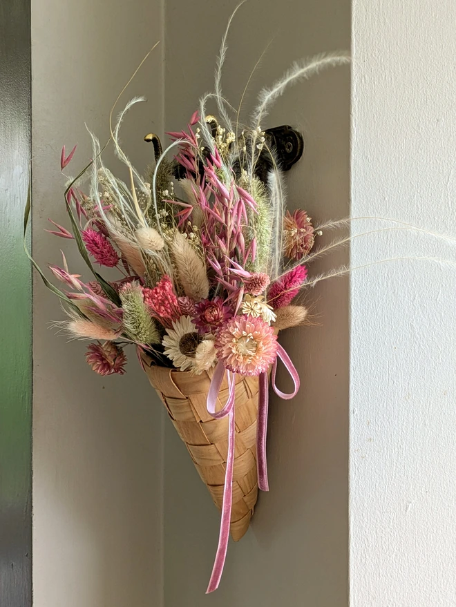 wooden braided cone basket with dried pink flowers hanging from a hook on a wall