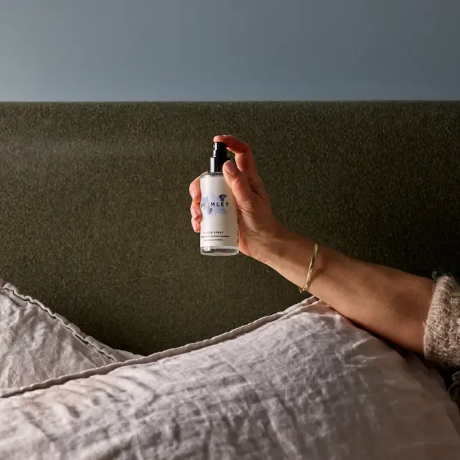 Hand holding a white spray bottle with black lid, finger placed on the spray pump. Against a grey headboard and resting on a light grey pillow.