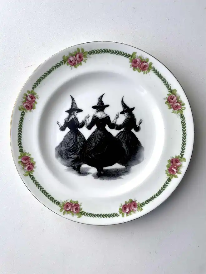 a coven of 3 witches dance hand in hand, the image is victorian in black and white and is printed directly onto an ornate vintage plate