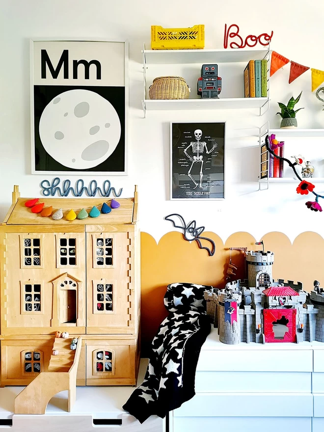 View of a childrens bedroom showing a chest of drawers with a wooden dollshouse and castle on top. A rolled up monochrome star blanket sits beside them, and above are black and white prints of a full moon and a skeleton. Wire word signs read 'Boo' and Shhhhhhhh' giving off Halloween vibes.gn