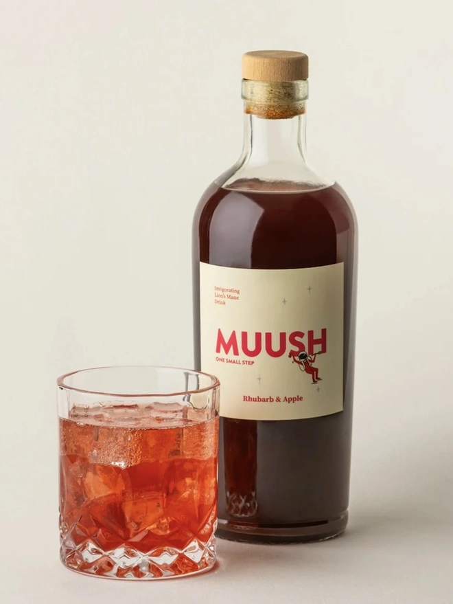 Rhubarb & Apple Elixir, a drink sitting in a glass next to a bottle of elixir on a plain surface. 