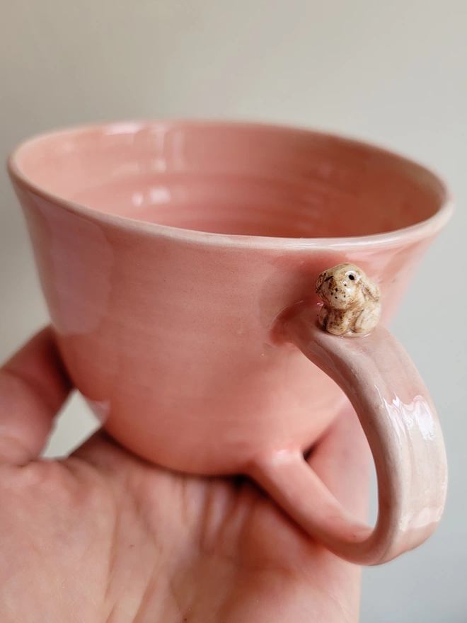 pink pottery tea cup with a tiny beige bunny rabbit on handle held in an outstretched hand 