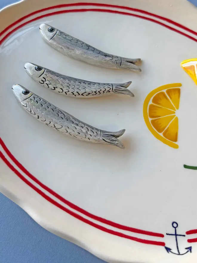 ceramic white platter with red border and anchor detail, with 3 ceramic sardines arranged on top, a ceramic wedge of lemon plus lemon painted on, with parsley detail, on a blue background