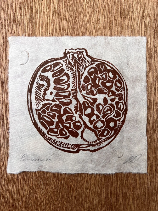 Hand-printed pomegranate lino print on natural handmade paper, featuring a bold botanical illustration of a ripe pomegranate.