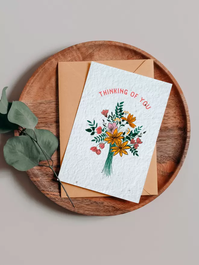 Seed Card with an illustration of a bouquet of flowers with ‘Thinking of You’ written above placed on a wooden tray next to a Eucalyptus branch