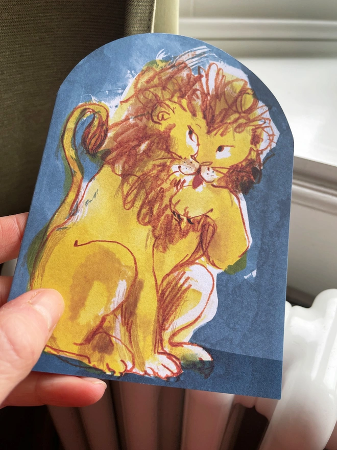 Esther Kent Lion illustrated arch-shaped greeting card, showing yellow and brown lion licking a paw, against a blue background.