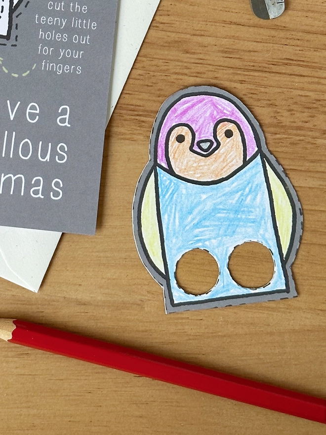 A colour in Christmas card with a penguin finger puppet design lays on a white envelope on a wooden desk.