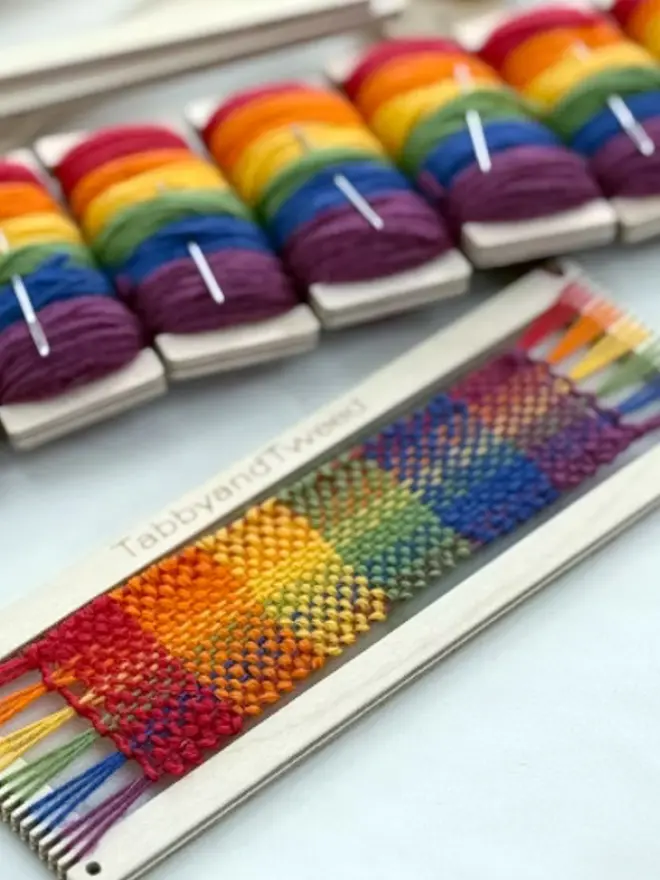 Rectangular beige loom woven with bright rainbow coloured yarns with spools of yarn in rainbow colours behind