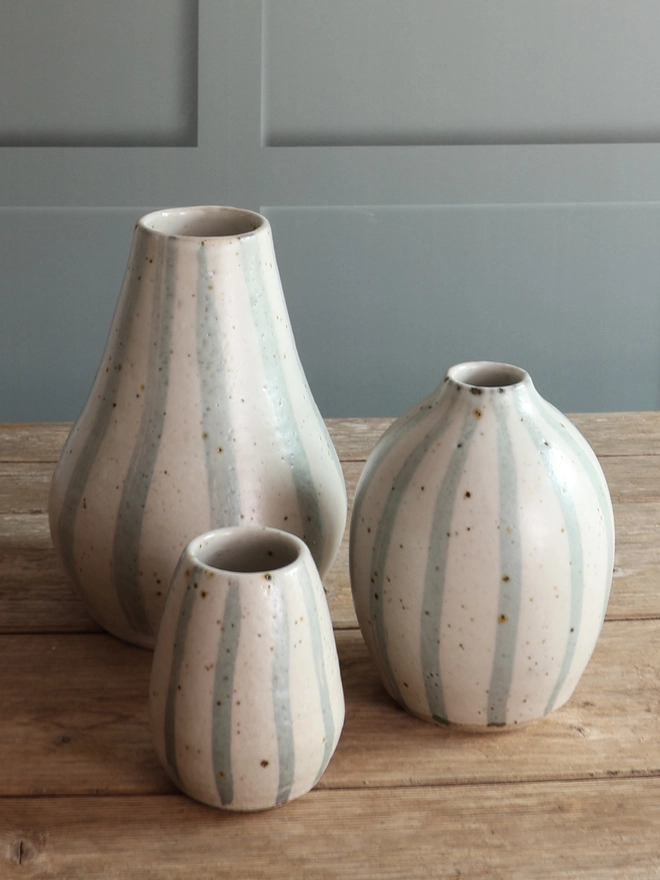 A collection of Handmade Ceramic Stoneware Vases in various sizes. Two of the vases feature a blue and white stripe design, and three of the vases feature a pink stripe design. The smallest Blue and White vases holds a bouquet of handmade ceramic flowers on copper wire stems.  