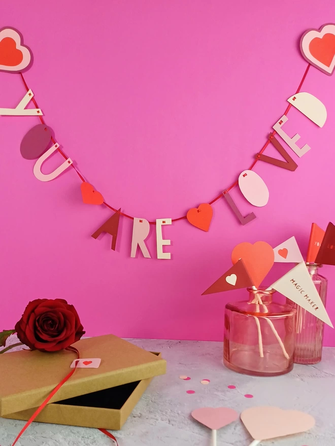 "You are loved" garland displayed on a bright pink wall. On a grey worktop a selection of appreciation flags and paper hearts are displayed in a pink vase