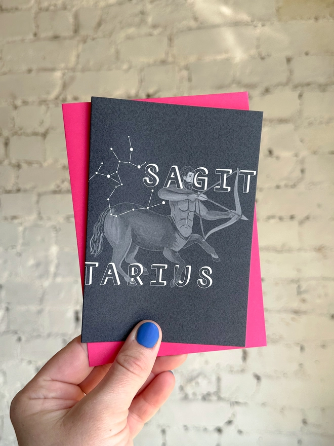 a greetings card featuring a greyscale illustration of a zodiac style centaur with the text “SAGITARIUS” with a neon pink envelope