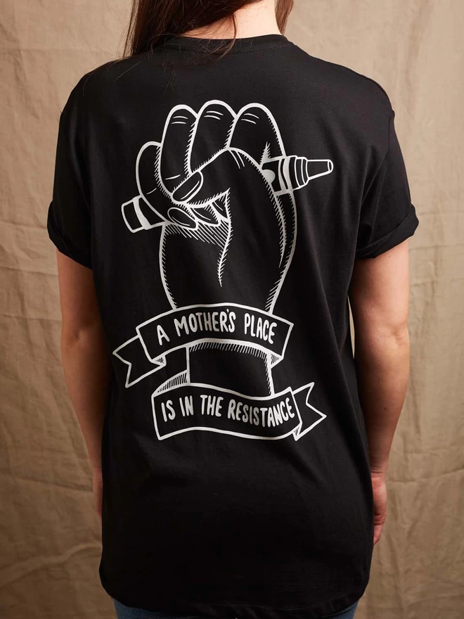Model is showing the back of the Black & Beech black A Mother’s Place is in the resistance t-shirt. The white print on the back shows a fist clutching a crayon and the with the words A Mother’s Place is in The Resistance written on a ribbon wrapped around the wrist 