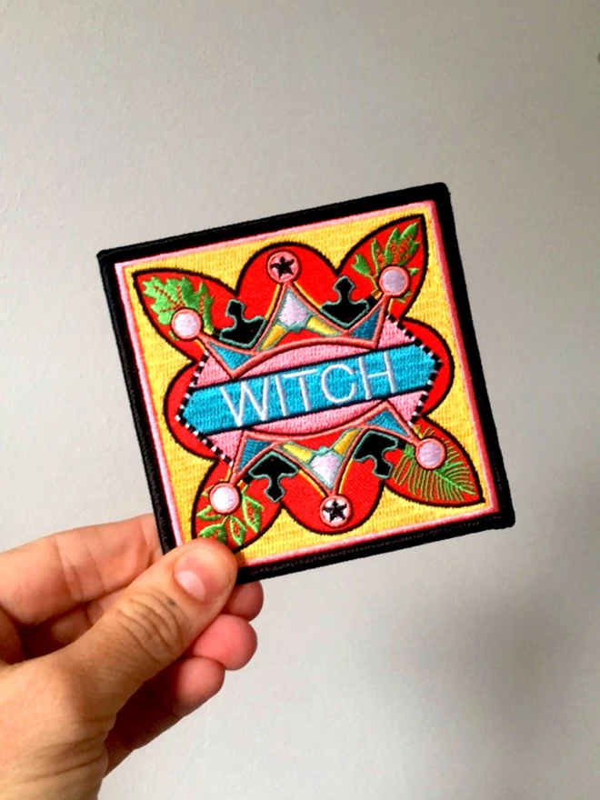A caucasian hand holds the fabric patch at the bottom left hand corner. In the centre of the patch the word “Witch” is embroidered in white on a light blue strip. Surrounding this is a colourful, bold design which sits on a yellow background, with a thin pink inner border and a thick black outer border.