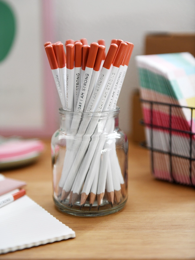 A glass jar full of white pencils with orange tips stands on a wooden desk surrounded by various stationery items.