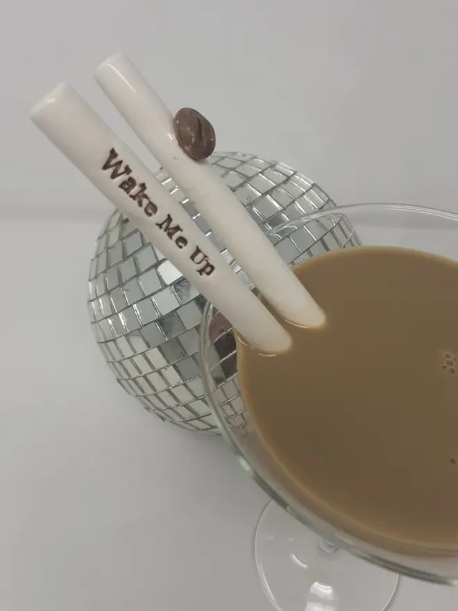 Two ceramic straws in a glass of Espresso Martini, One straw has the words "Wake Me Up" in coffee brown and the other is decorated with a coffee bean. There is a mirror ball in the background.