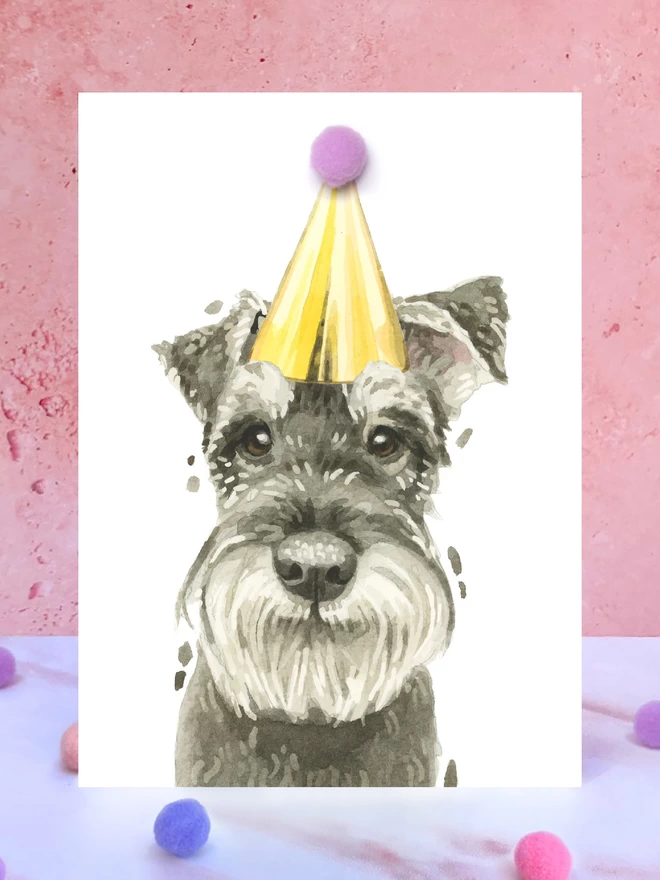 A greeting card featuring a hand painted design of a schnauzer, stood upright on a marble surface surrounded by pompoms. 