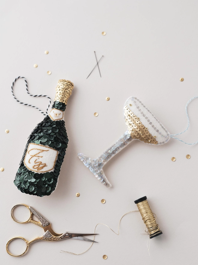 A green champagne bottle ornament on a cream table with a champagne coupe ornament next to it