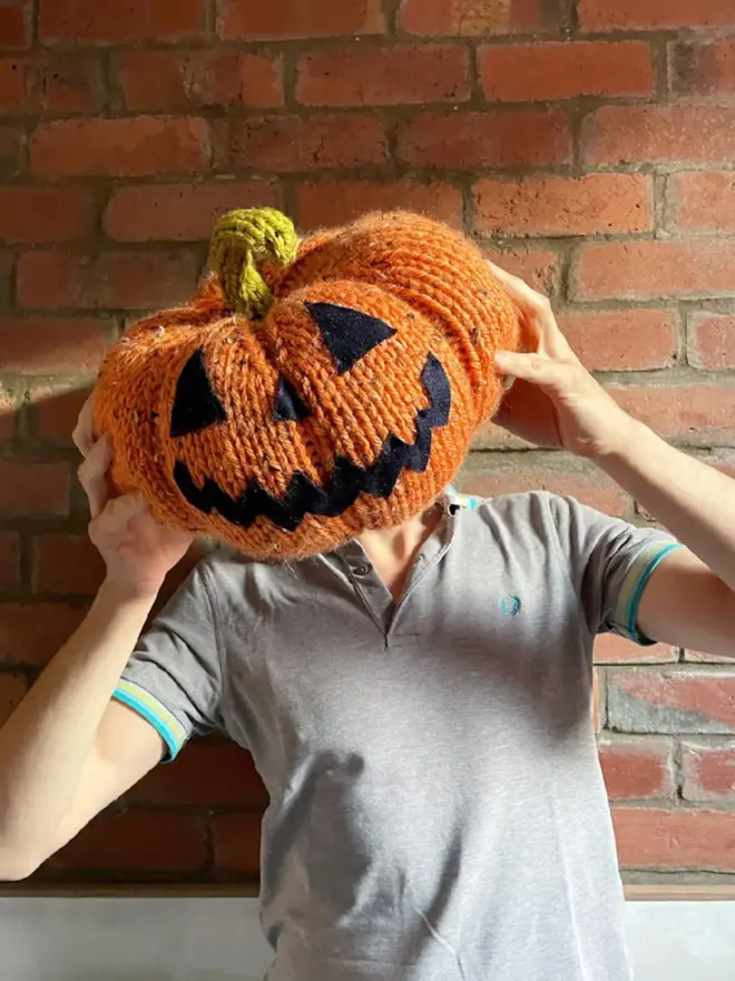person in grey shirt, holding large knitted jack o lantern in front of their face
