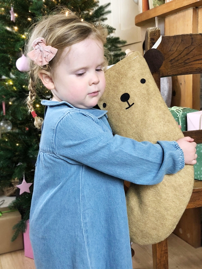 A young girl wearing a denim dress stands in front of a Christmas tree hugging a handmade felt bear stocking.